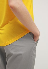 Personne portant une chemise jaune, la main droite dans la poche d'un pantalon gris clair, sur un fond uni.