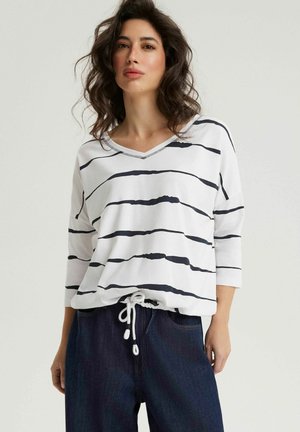 Kobieta o falowanych włosach, ubrana w biały T-shirt z dekoltem w serek z nieregularnymi czarnymi paskami i granatowe spodnie ze ściągaczem na jednolitym tle.