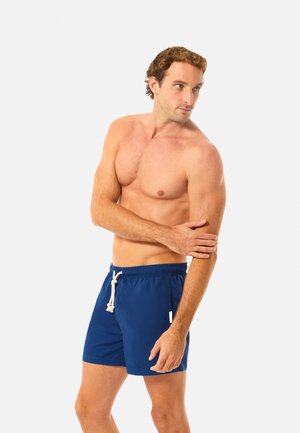 Mies, jolla on lyhyet ruskeat hiukset ja hänellä on siniset uimashortsit, joissa valkoinen kiristysnauha. Hän seisoo ja pitää vasenta kättään samalla kun katsoo oikealle.