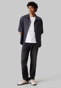 Korte mouw donkergrijze overhemden over een wit T-shirt, gecombineerd met zwarte jeans en beige sportschoenen. Eenvoudig ontwerp, relaxte pasvorm.
