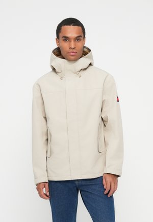 Hombre vestido con una chaqueta beige con capucha y bolsillos delanteros, y jeans azules, de pie contra un fondo blanco liso.