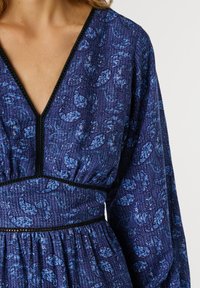 Donna che indossa un vestito blu con motivo paisley, scollo a V profondo e maniche lunghe, con dettagli di rifinitura neri sul collo e in vita.
