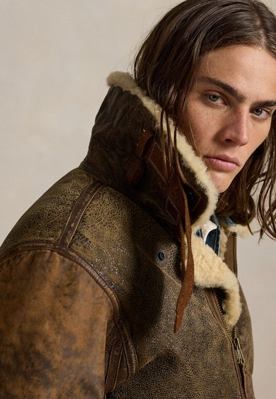 Giovane con capelli lunghi e castani che indossa una giacca di pelle marrone in shearling con colletto alto e che guarda leggermente lontano dalla fotocamera.
