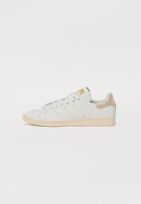 Zapatillas blancas de cuero con detalles perforados en los laterales, acento en ante beige en el talón, cordones planos y suela de color claro. Logo dorado en la lengüeta.
