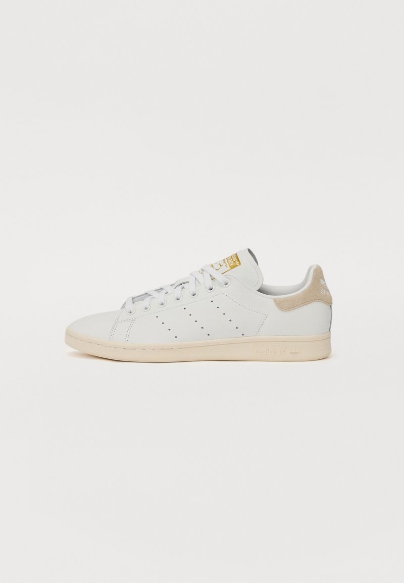 Zapatillas blancas de cuero con detalles perforados en los laterales, acento en ante beige en el talón, cordones planos y suela de color claro. Logo dorado en la lengüeta.