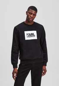 Sweatshirt negru cu mansete ribbatute, decolteu rotund și un logo dreptunghiular alb cu "KARL LAGERFELD" scris cu litere îndrăznețe negre.
