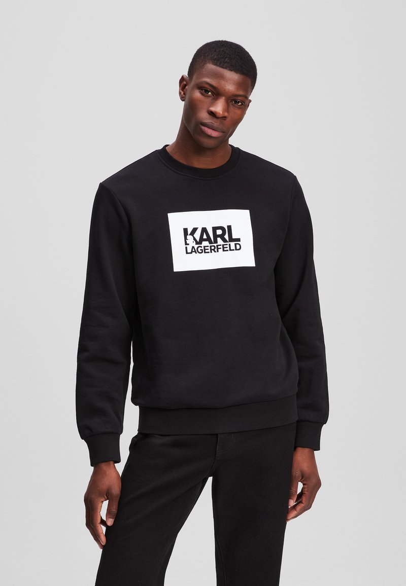 Sweatshirt negru cu mansete ribbatute, decolteu rotund și un logo dreptunghiular alb cu "KARL LAGERFELD" scris cu litere îndrăznețe negre.