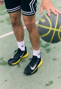 Black and yellow athletic shoes with a textured upper, white swoosh logo, paired with white socks on a green sports court.