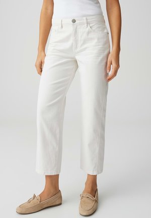Vrouw draagt witte rechtvallende cropped jeans, witte tanktop en beige suède loafers, staand tegen een effen achtergrond.