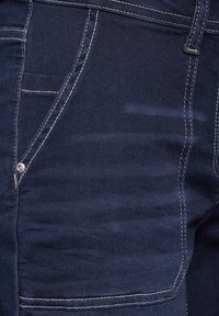 Jeans in denim blu scuro con cuciture chiare in contrasto, dotati di una tasca frontale con rivetto metallico e una finitura liscia e testurizzata.