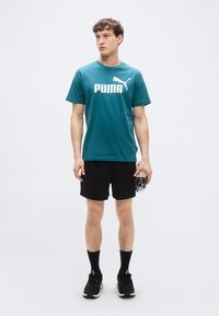 Τιρκουάζ βαμβακερό μπλουζάκι με λευκό λογότυπο "PUMA" και γραφικό πούμα. Μαύρο σορτς, μαύρες κάλτσες και μαύρα αθλητικά παπούτσια. Κρατάει ένα μπουκάλι νερό.