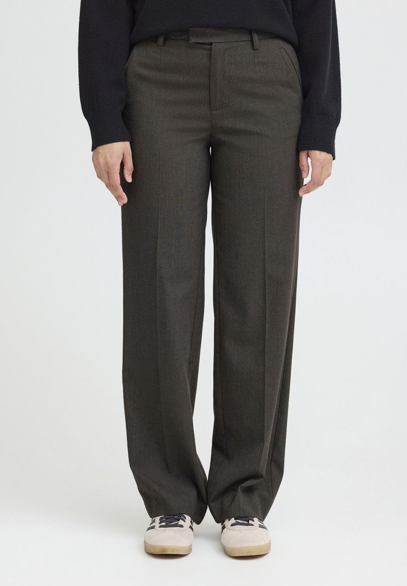 Personne portant un pantalon vert foncé à jambes larges et des baskets blanches, debout devant un fond clair uni.
