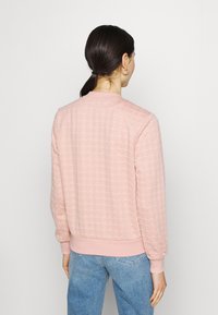 Donna con i capelli scuri raccolti in una coda di cavallo, che indossa un maglione leggero rosa chiaro dalla texture e jeans blu, in piedi di spalle su uno sfondo bianco.