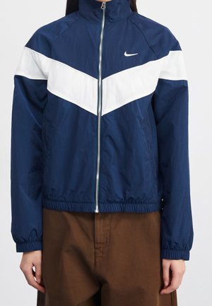 Osoba nosící tmavě modrou bundu na zip s bílým šikmým pruhem a logem Nike, kombinovanou s hnědými kalhotami.