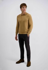 Beige gerippter Strickpullover mit Drei-Knopf-Leiste, kombiniert mit dunkelgrauen Hosen und braunen suedeboots, vor einem neutralen Hintergrund.