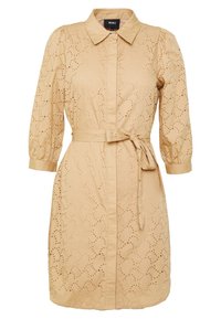 Vestido beige con cuello camisero, cierre de botones y cinturón en la cintura. Presenta un patrón de broderie anglaise y mangas abullonadas de tres cuartos.