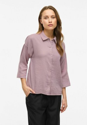 Chemise rose clair à boutons, en tissu doux et texturé, avec un col et des manches trois-quarts, assortie à un pantalon noir ample.