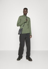 Maglione a collo alto verde a maglia, pantaloni neri a gamba larga, borsa nera a tracolla, scarpe nere stringate con suole spesse, sfondo semplice.