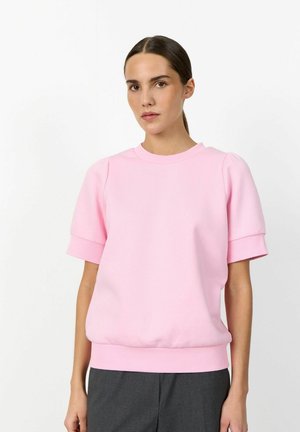 Žena s tmavými vlasmi zviazanými do nízkeho copu, oblečená v svetloružovej mikine s krátkym rukávom a tmavosivých nohaviciach na bielom pozadí.