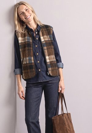Smilende kvinde læner sig op ad en væg iført ternet vest over en denimskjorte og jeans, mens hun holder en brun taske i håndtagene.