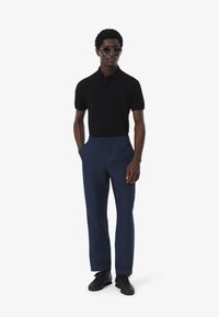 Camisa polo negra de mangas cortas, pantalones azul marino con cinturilla elástica y zapatillas negras. Los accesorios incluyen gafas de sol.