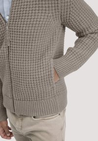 Hellgraue gestrickte Zip-up-Cardigan mit einem strukturierten Muster, gerippten Bündchen und zwei Seitentaschen. Das Material wirkt dick und kuschelig.