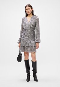 Hõbedane sädelev pakkekleit pikkade varrukatega ja kohandatud taljega, rippuvate äärtega ja kombineeritud põlvini ulatuvate mustade saapade ning väikese kotiga.