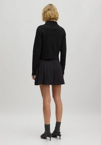 Schwarzer, cropped Pullover mit geripptem Kragen, kombiniert mit einem gefältelten schwarzen Rock. Das Modell trägt dunkle Socken und schwarze Absätze und zeigt ein minimalistisches Design.