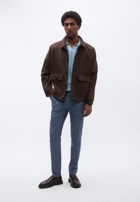 Καφέ σακάκι με κουμπιά και δύο μπροστινές τσέπες πάνω από ένα γαλάζιο πλεκτό πουλόβερ, συνδυασμένο με μπλε παντελόνια και σκούρα παπούτσια.