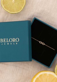 Goldenes Unendlichkeit-Armband in einer schwarz samtierten teal-farbenen Schachtel mit goldener Schrift, umgeben von Limettenscheiben auf einem strukturierten beigen Hintergrund.