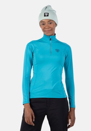 Langarm, turquoise prestatie shirt met een halvemantel kraag, gecombineerd met een lichtblauwe beanie met een zwart logo patch.
