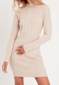 Abito maglione in beige chiaro con motivi a treccia, collo alto e maniche lunghe. Forma aderente con polsini e orlo a coste.