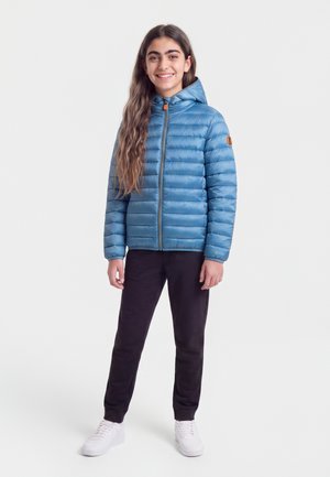 Ung person med langt hår iført en blå dunjakke, sorte bukser og hvide sneakers, der står og smiler mod en ensfarvet baggrund.