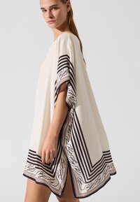 Bijeli kaftan s prugastim smeđim obrubom i uzorkovanim detaljima. Labav kroj, kratki rukavi i bočni razrezi za lakše kretanje. Glatka tekstura.