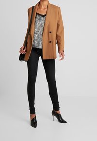 Blazer marrón de doble botonadura sobre una blusa de patrón negro y plata, combinado con jeans ajustados negros y tacones puntiagudos negros.