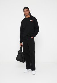 Sudadera negra con un logo colorido, combinada con pantalones negros. El modelo sostiene un bolso negro y lleva zapatillas blancas. Fondo sencillo.