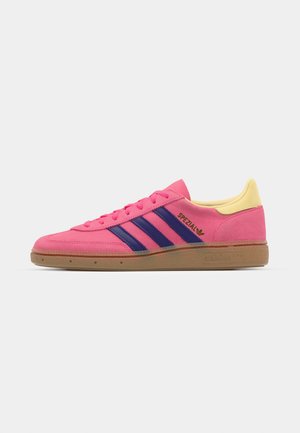 Różowe zamszowe sneakersy z żółtym piętkiem, z trzema niebieskimi paskami i gumową podeszwą. Logo "Spezial" jest nadrukowane na czarno z boku.