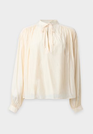 Mujer de pie vestida con blusa crema de manga larga con lazo en el cuello, vaqueros oscuros de tiro alto y corte recto, y zapatos negros puntiagudos sobre fondo blanco.
