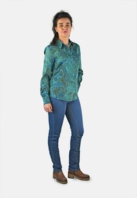 Camisa de botones de seda en color verde azulado con un patrón de paisley, de mangas largas, combinada con jeans azules y botas cortas marrón, de pie sobre un fondo blanco.