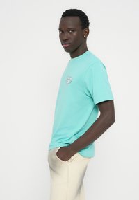 Τυρκουάζ βαμβακερό μπλουζάκι με κοντά μανίκια και στρογγυλή λαιμόκοψη, με μικρό γραφικό σε λευκό και μπλε στο στήθος. Φορεμένο με ανοιχτόχρωμο σορτς.