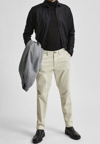 Černá košile na knoflíky přes černý rolák, krémové chinos kalhoty, šedá mikina přes ruku a černé kotníkové boty. Hladké textury a neutrální barvy.