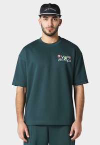 Mørkegrønn t-skjorte med korte ermer og avslappet passform, med blomstrete "PAS DE MONACO" broderi foran og strukturgarn.