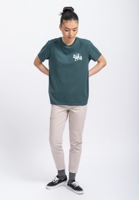 Teal katoenen t-shirt met een kleine witte grafiek op de linkerborst, gecombineerd met lichtbeige tapered broeken en zwarte sneakers.