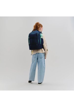 Junge Person mit roten Haaren, die eine beige Jacke, weite blaue Jeans, schwarze Turnschuhe und einen großen marineblauen Rucksack mit türkisfarbenen Akzenten trägt.