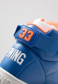 Baskets en cuir bleu avec "33" orange sur la sangle et texte blanc "EWING" au talon, avec semelle blanche et col matelassé orange.