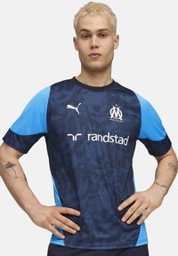 Maillot à manches courtes bleu et marine avec un motif à pois, comportant le logo Puma, l'emblème de Marseille et le texte "randstad" en blanc.