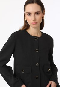 Veste noire avec un col rond, à manches longues et boutons circulaires dorés. Possède deux poches avant avec fermetures à rabat. Texture lisse.