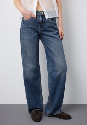 Persona che indossa jeans blu larghi con la mano sinistra in tasca, abbinati a mocassini marroni e una camicia bianca trasparente.