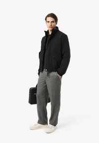 Man med svart jacka, grå byxor, vita sneakers, håller en svart portfölj, står mot vit bakgrund, vänd mot kameran.