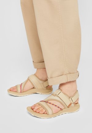Voeten met beige sandalen met verstelbare bandjes en kleurrijke gevlochten details, gecombineerd met opgerolde lichtbeige broekspijpen tegen een neutrale achtergrond.
